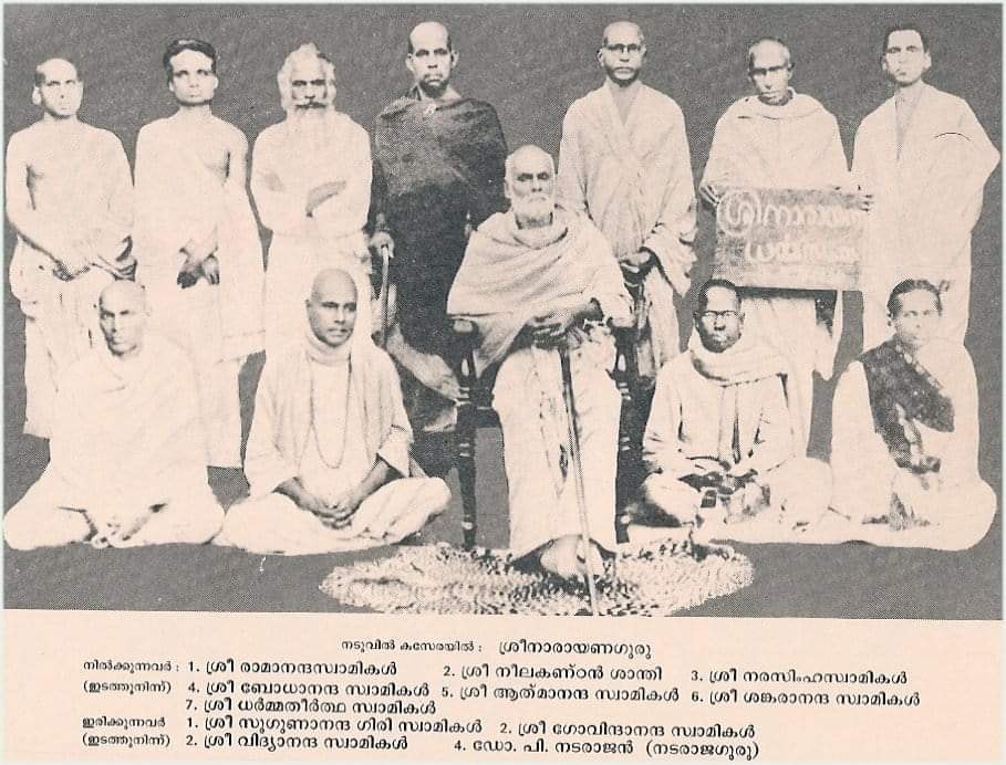 A group photograph of eleven individuals, predominantly dressed in traditional attire, posed together. The front row features several seated figures, while the back row stands behind them. One individual is holding a sign with text written in Malayalam.