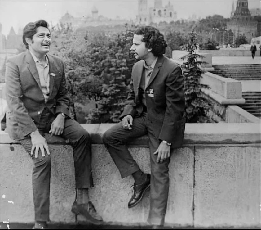 Black and white photo of two men sitting on a stone wall, engaged in conversation, with trees and buildings in the background.