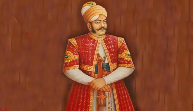A portrait of a royal figure wearing a traditional red and gold embellished attire, with a turban, standing confidently against a brown background.