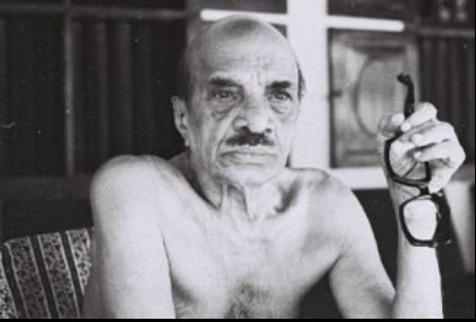 Black and white photograph of an older man sitting indoors, shirtless, with a serious expression, holding a pair of glasses in his right hand.