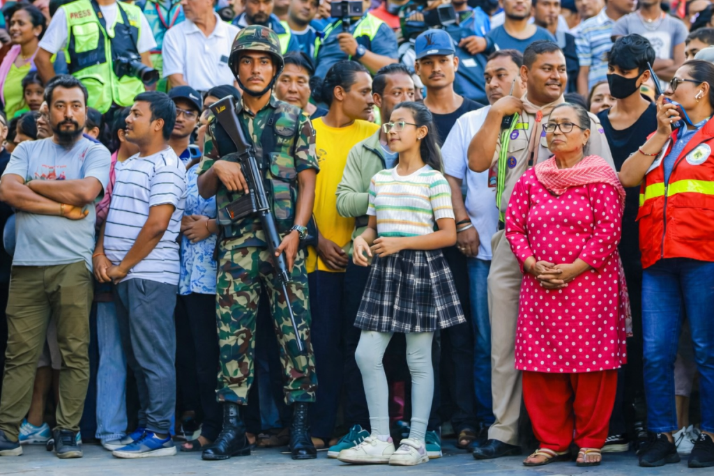 A large crowd of people gathered outdoors, including a soldier in camouflage with a rifle, a young girl in a striped shirt and plaid skirt, and a woman in a pink outfit with polka dots. There are various expressions among the crowd, and a few individuals are holding cameras.