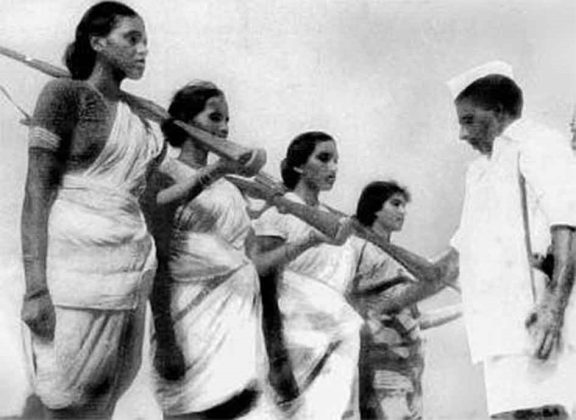 A historical black and white photograph depicting a group of women dressed in traditional attire carrying wooden staffs, standing in conversation with a man in a white uniform and cap.