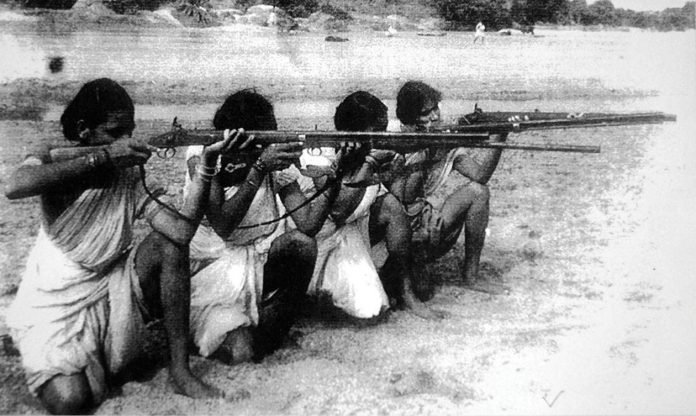 A historic black and white photograph showing four individuals in traditional clothing aiming rifles while seated on the ground, set against a backdrop of a sandy landscape.