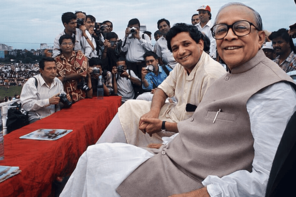 Two men seated at a table with a red cloth, smiling at the camera, while a crowd of photographers and onlookers is in the background.