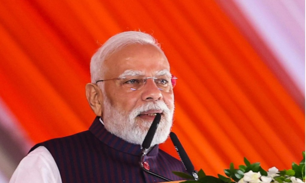 A man with a white beard and glasses, dressed in a traditional dark outfit, speaking at an event with an orange and white background.
