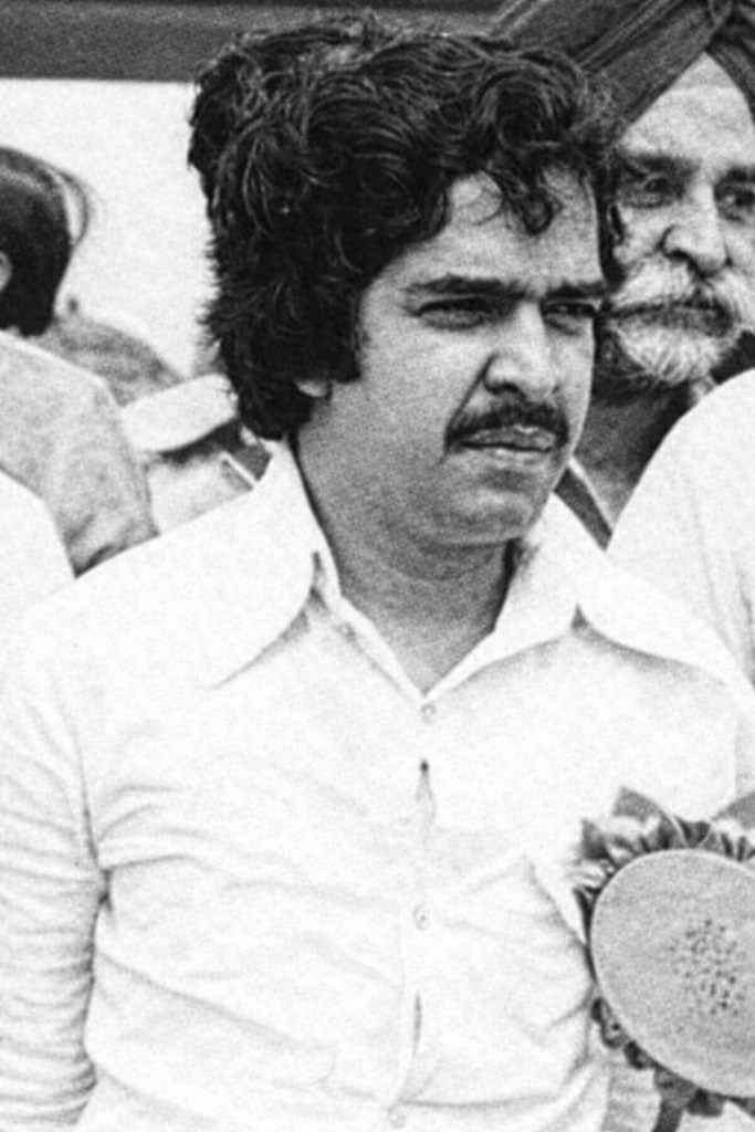 A young man with dark, curly hair and a moustache, wearing a light-coloured shirt, stands in a candid outdoor setting. He appears to be engaged in conversation, with a serious expression on his face. In the background, other individuals are partially visible.