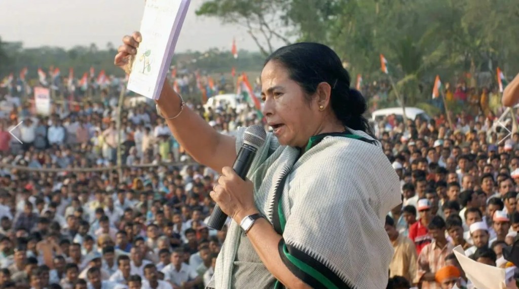 A woman holding a book and microphone speaks passionately to a large crowd at a rally, surrounded by flags and banners.